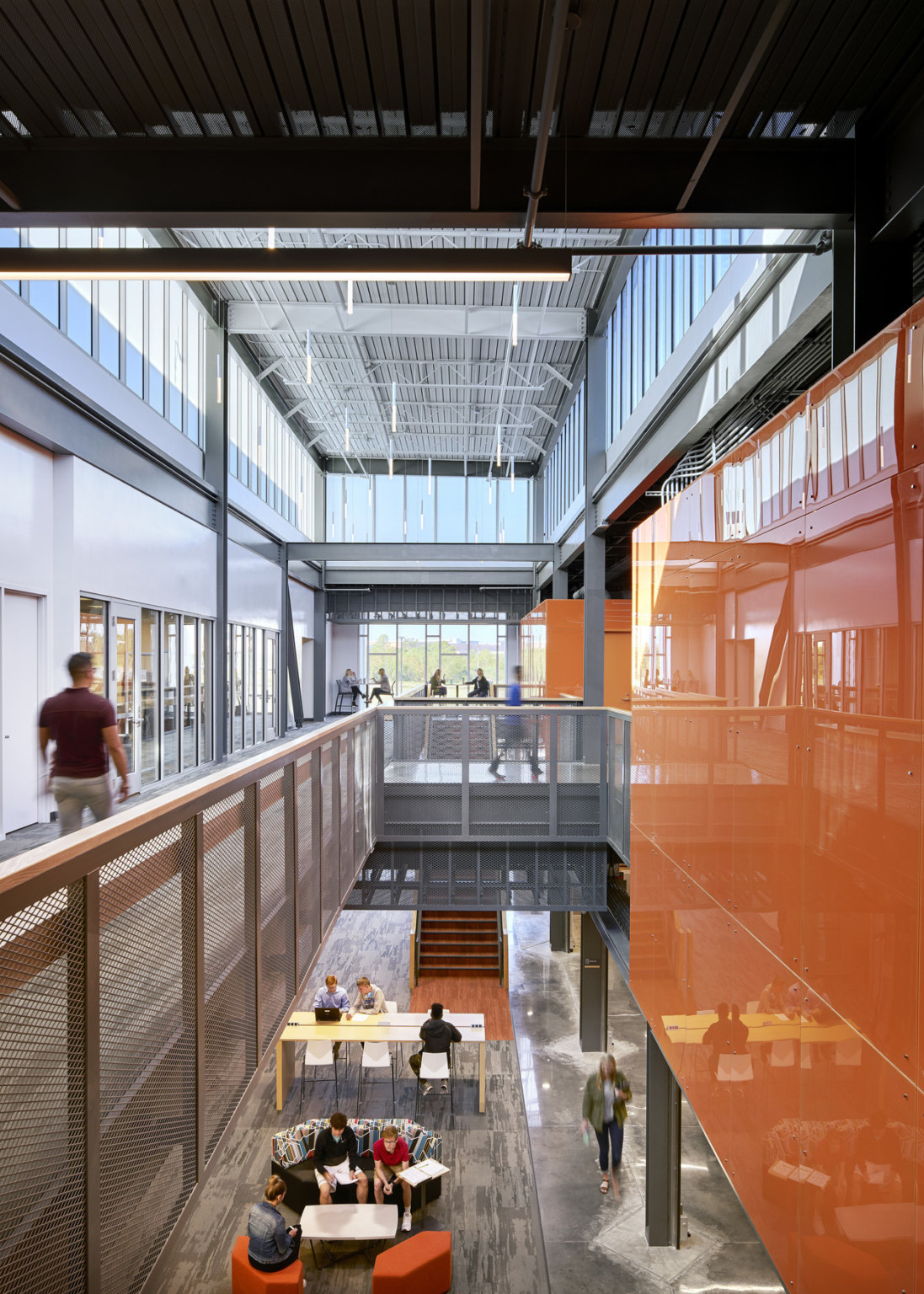 View of atrium from 2nd floor walkway ending in double height windows. Orange walls on the right, and a seating area below