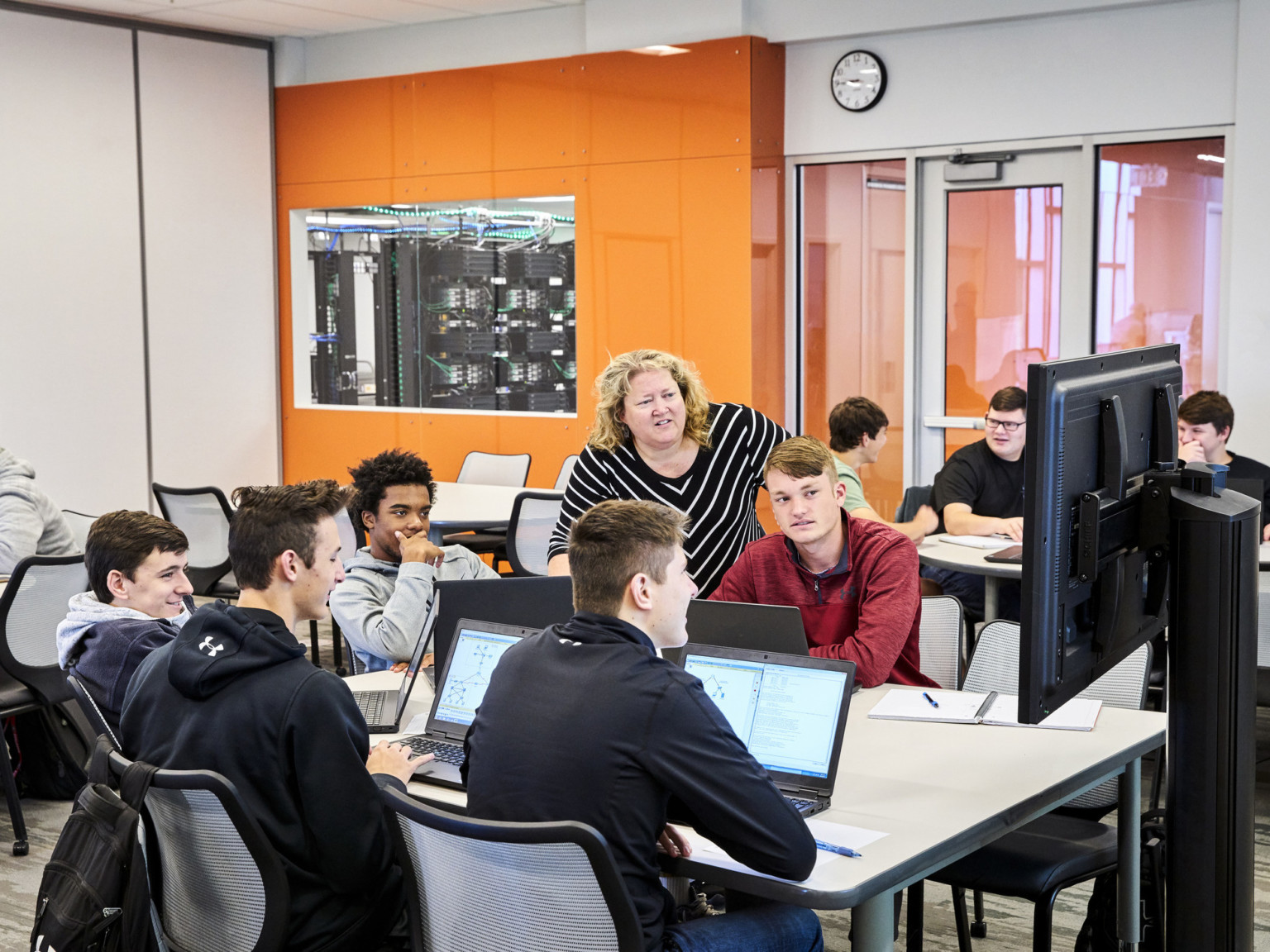 In a white classroom with an orange accent wall, students at a table look at a portable screen with their teacher