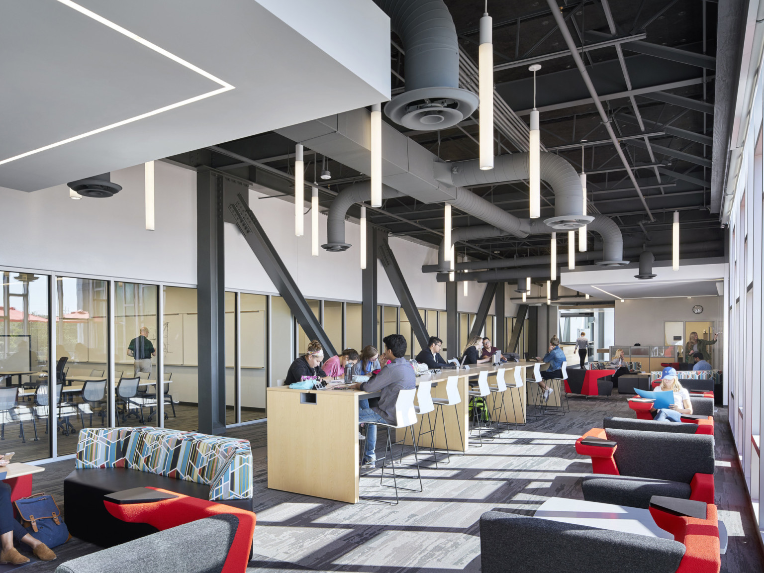 Hallway with seating spaces. Floor to ceiling windows, right, with transparent wall classrooms left, and drop ceiling details