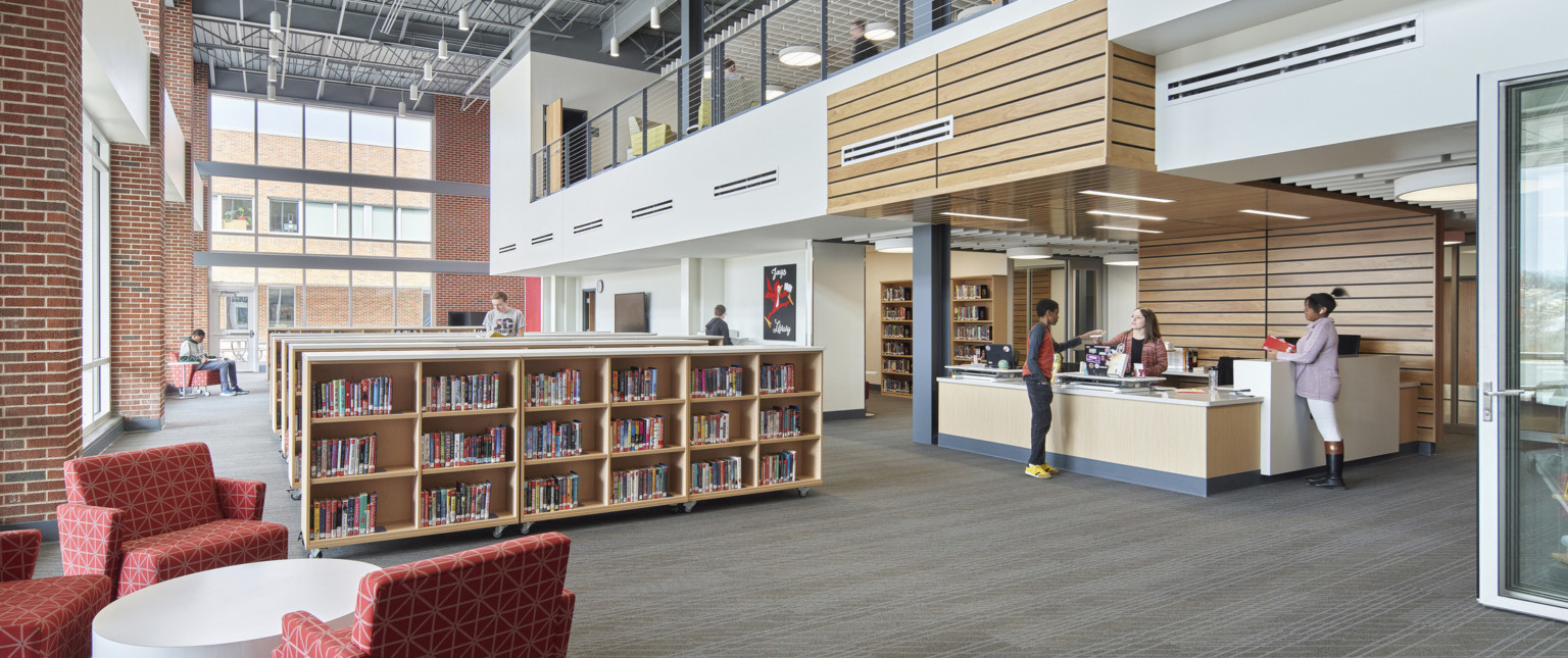 Half height bookcases in library. Double height windows wrapping from left wall to back. Balcony overhang above desk, right