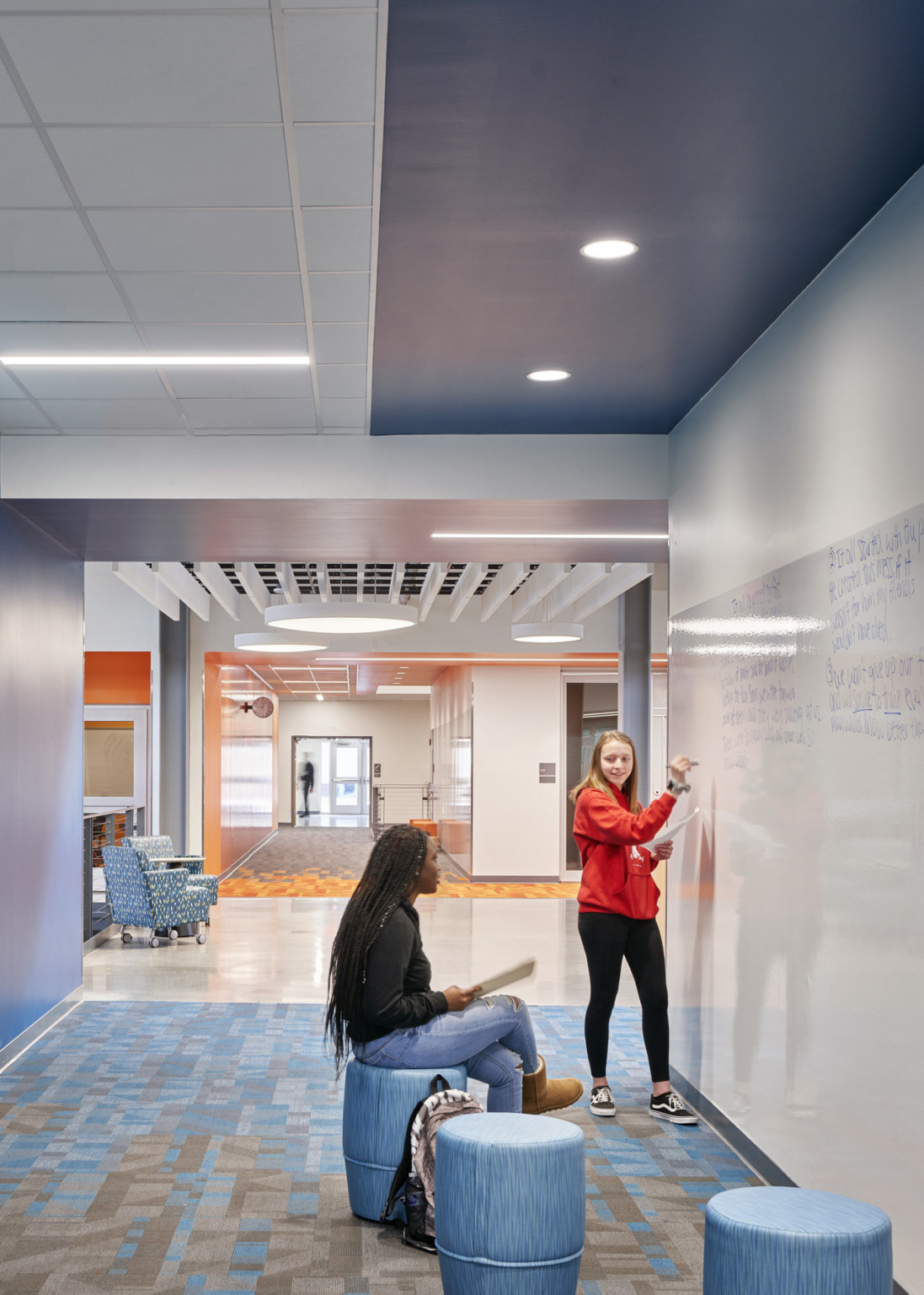 White hallway with blue stools around whiteboard. Hallway connects to seating near stairwell below slat ceiling detail