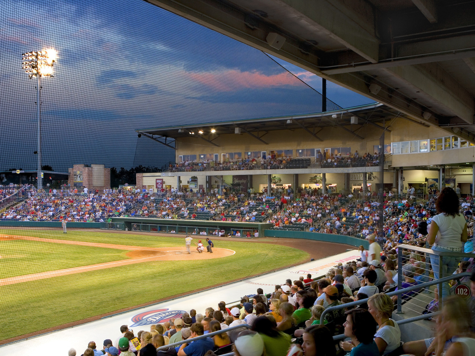 Lower deck seating along third base line with suites above. View through net of batter at home plate and suites opposite