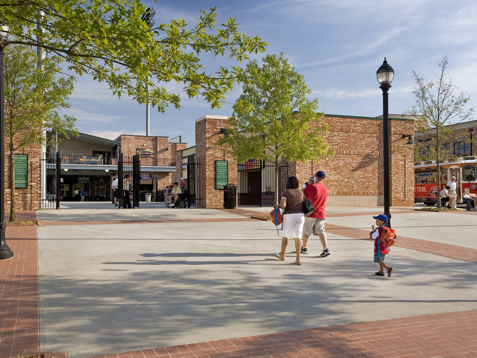 Tree and brick lined plaza leading to entry gate of stadium. Brick wall sections connected by black metal fence