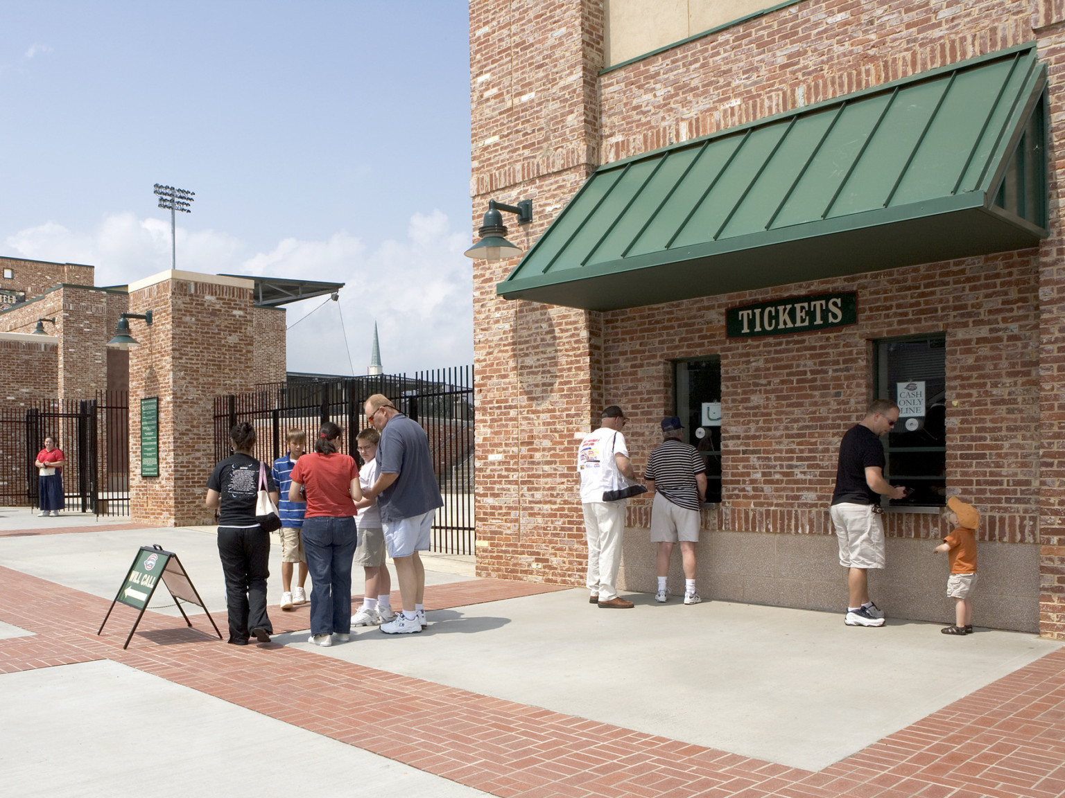 Brick building with 2 ticket windows sheltered by green awning to the right side of black fence and gated entrance