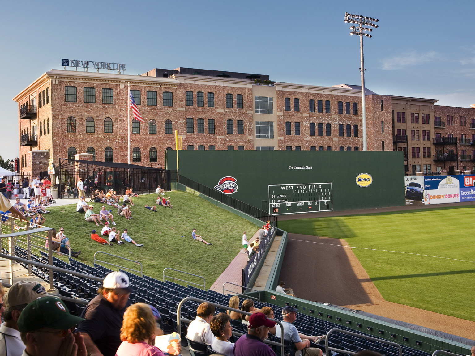Lawn seating on sloped hill beyond third base foul line. Right, a green wall with score board in front of a brick building