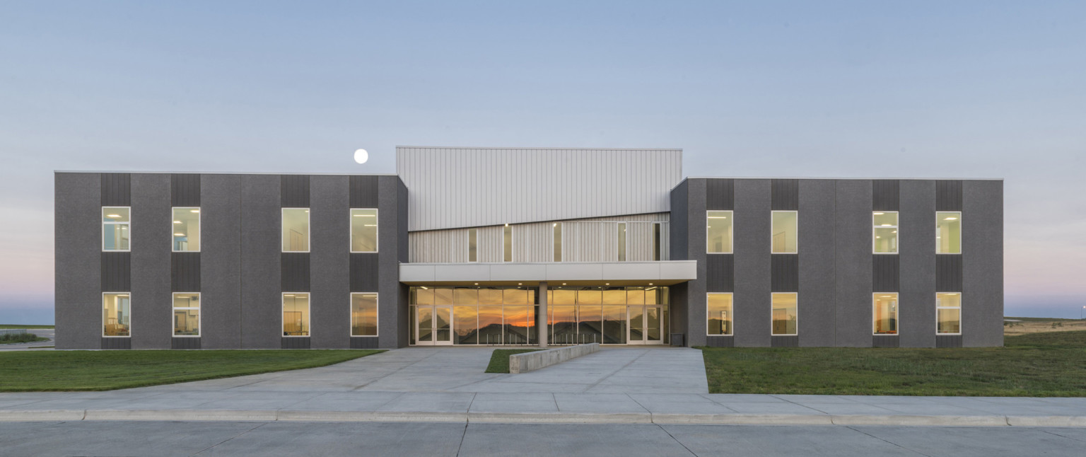 Dark grey building sections frame recessed center with glass base and canopy below silver panel 2nd floor. Driveway to front