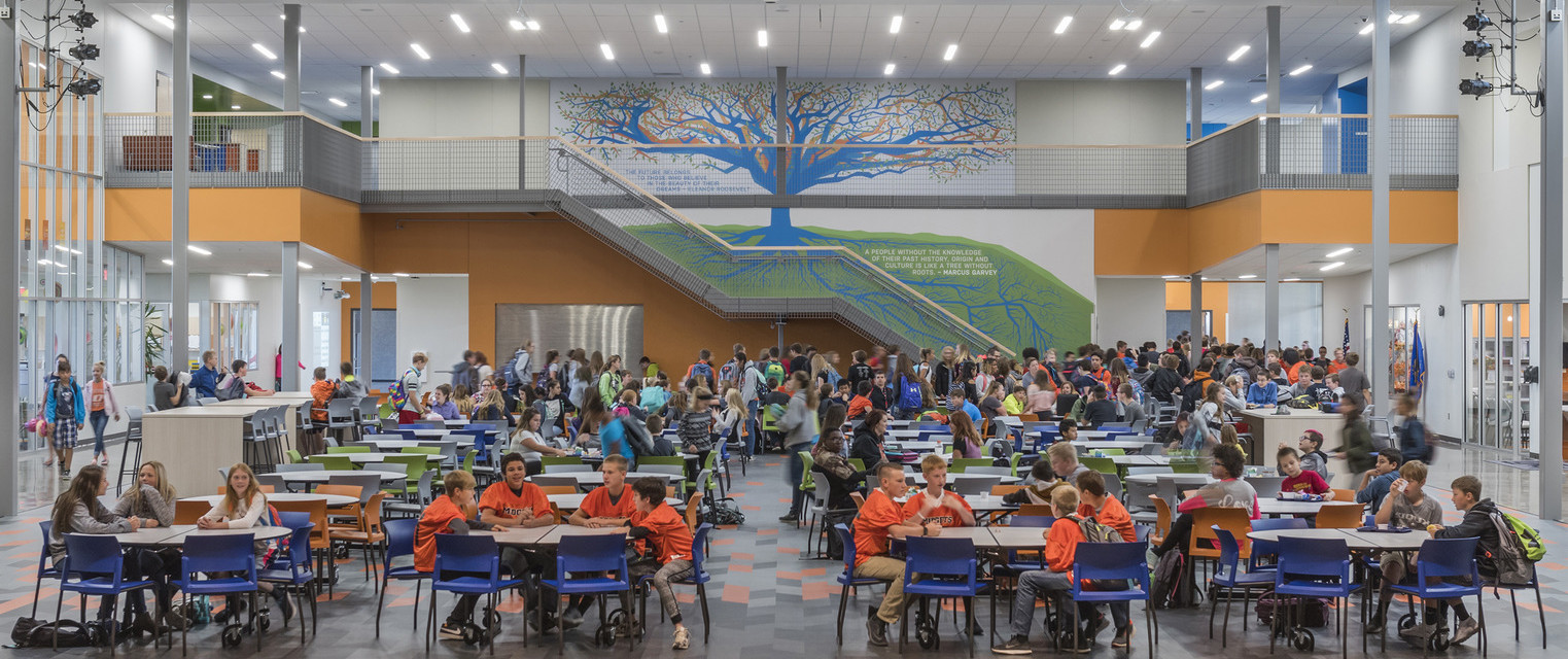 Double height room with tables and chairs on 1st floor. Orange stairs at back wall with abstract blue and orange tree mural