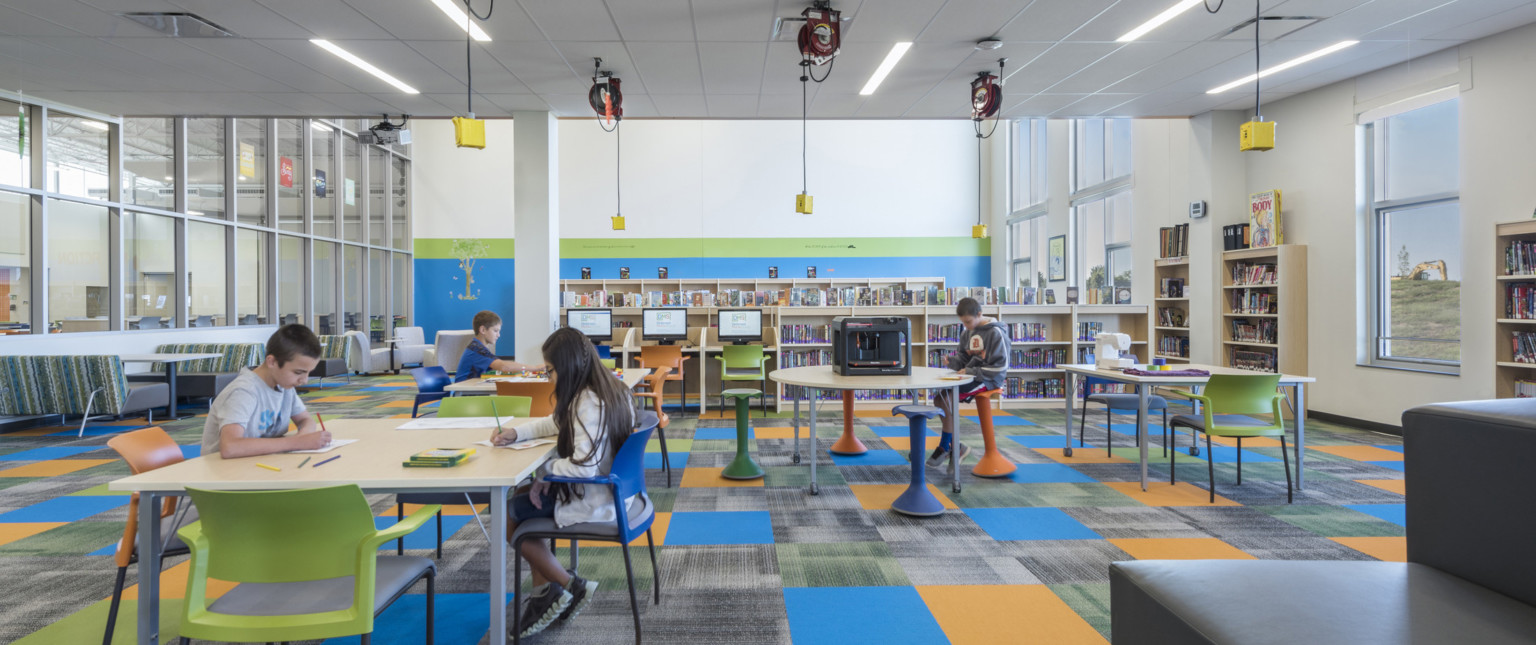 Flexible seating space in carpeted room with floor to ceiling windows to hall, right, and rows of half height book shelves