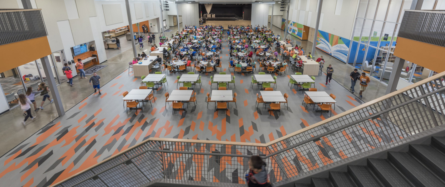 View from stairs down to cafeteria tables. Book and music murals on right by library. Cafeteria counters and sink, left