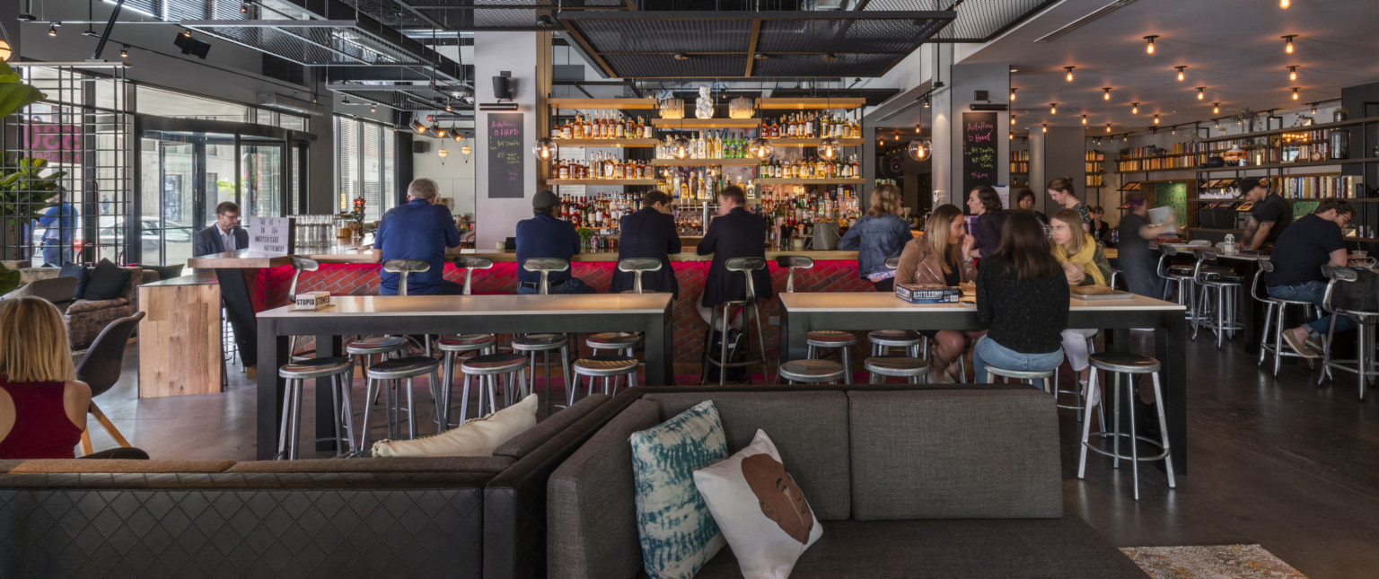 Red brick bar with wood counter and behind the bar shelving, high top tables and stools sit between bar and couches