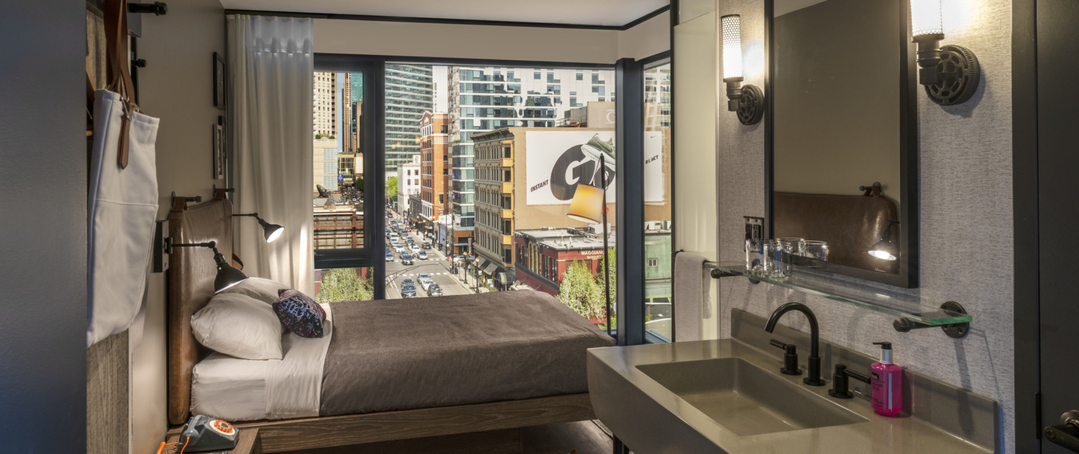 View of guest room from grey sink, looking towards bed and large corner window, white room with grey around sink and mirror