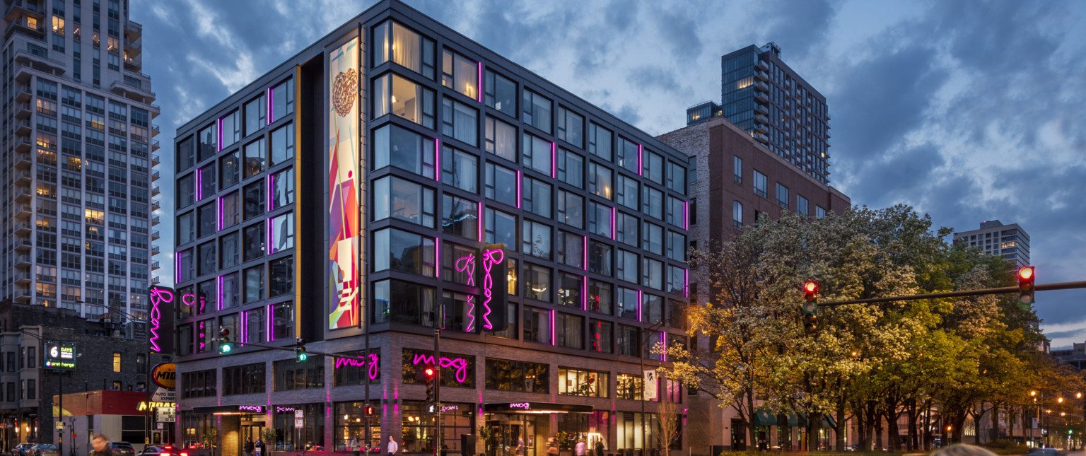 Corner view of the Moxy hotel with illuminated mural and pink lights at various windows, 2 brick stories, exposed steel above
