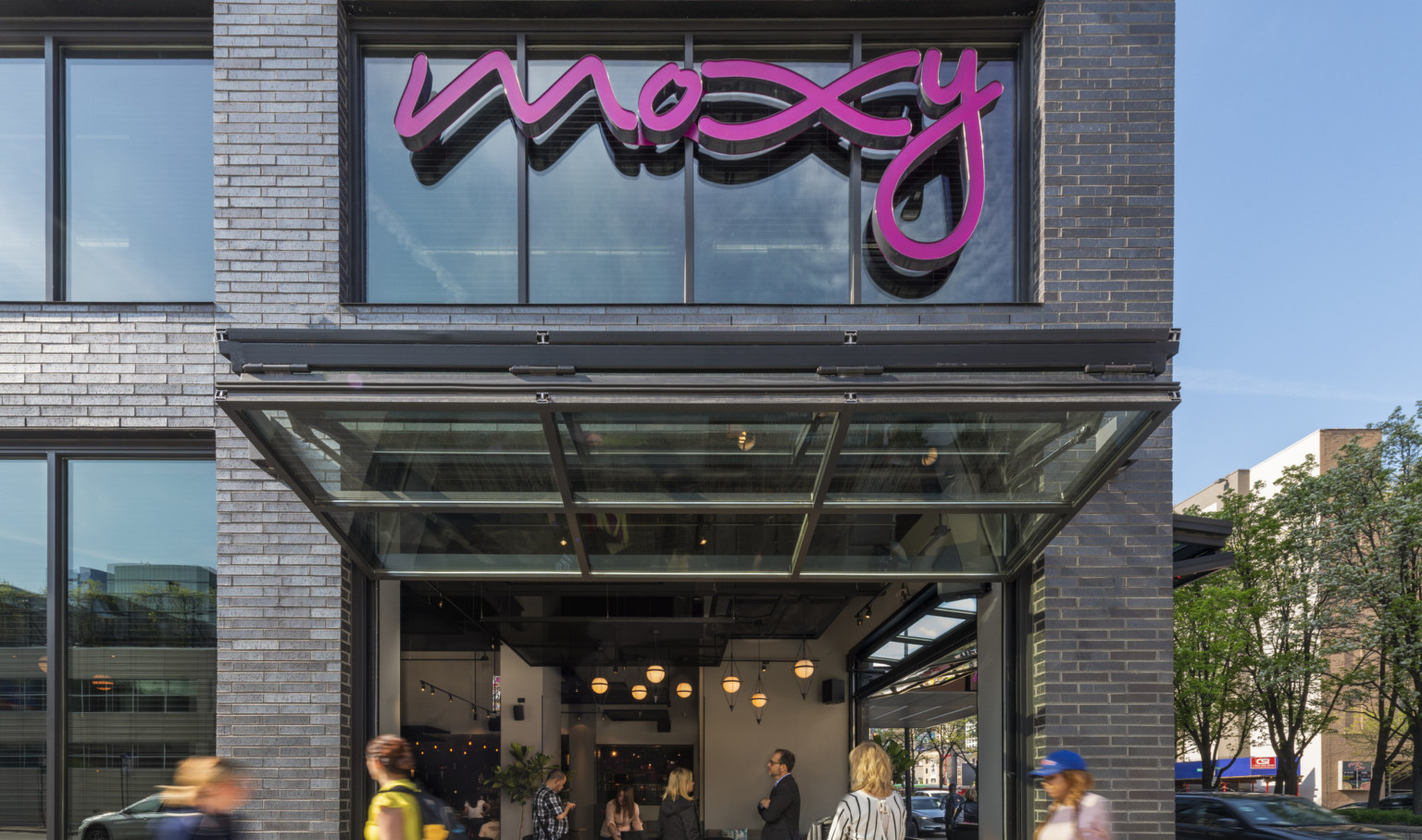 1st floor corner of building with window below pink Moxy sign opened up and out above sidewalk looking into dining area