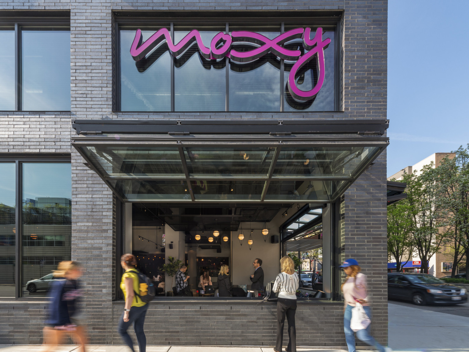 1st floor corner of building with window below pink Moxy sign opened up and out above sidewalk looking into dining area