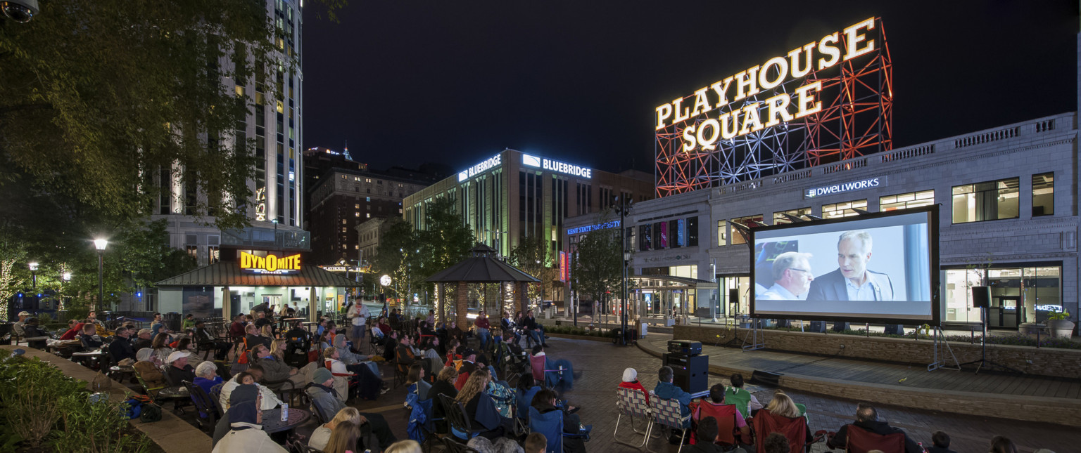 Audience sits on the street watching an outdoor movie on a screen. A Playhouse Square sign is illuminated in the background