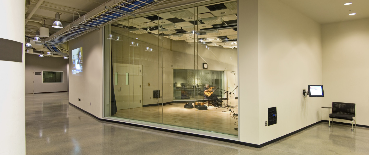 Guitarist in room with partial glass walls, hard wood flooring, and acoustic panels on ceiling. Outside room is a hall