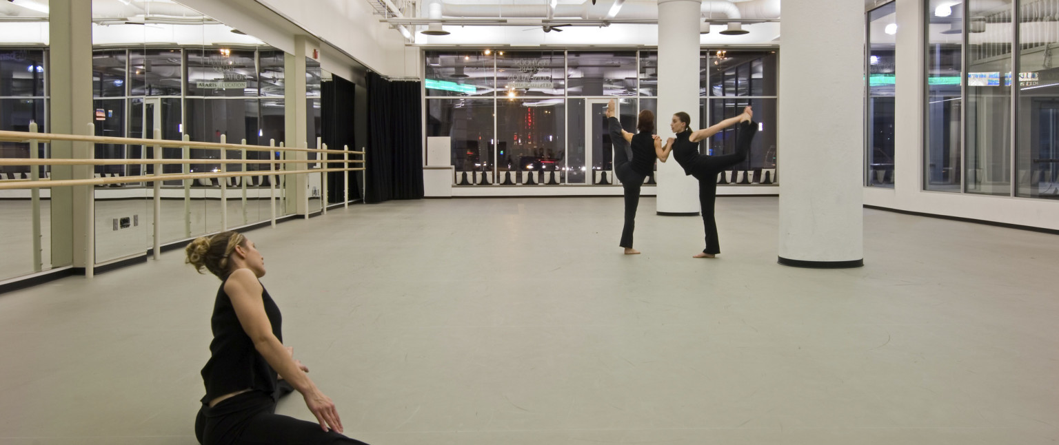 Three dancers in a white studio with large window walls looking out in the city. Left wall is mirrored and has a barre