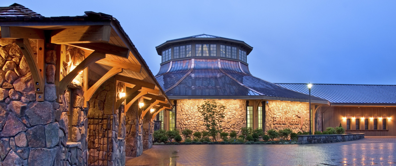 Stone gate with wooden roof and trusses next to round stone building with metal roof and a band of windows near the top