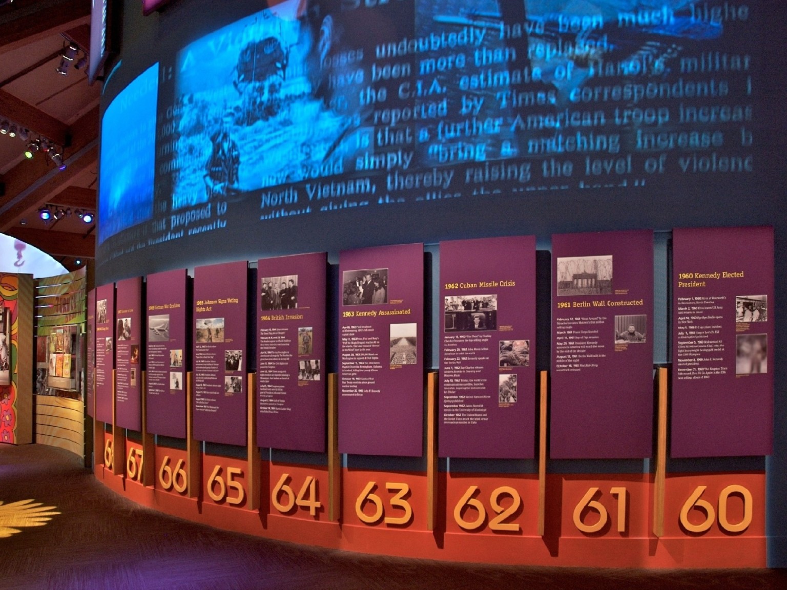A wall with timeline and display panels of information curves beneath a video display; museum at bethel woods