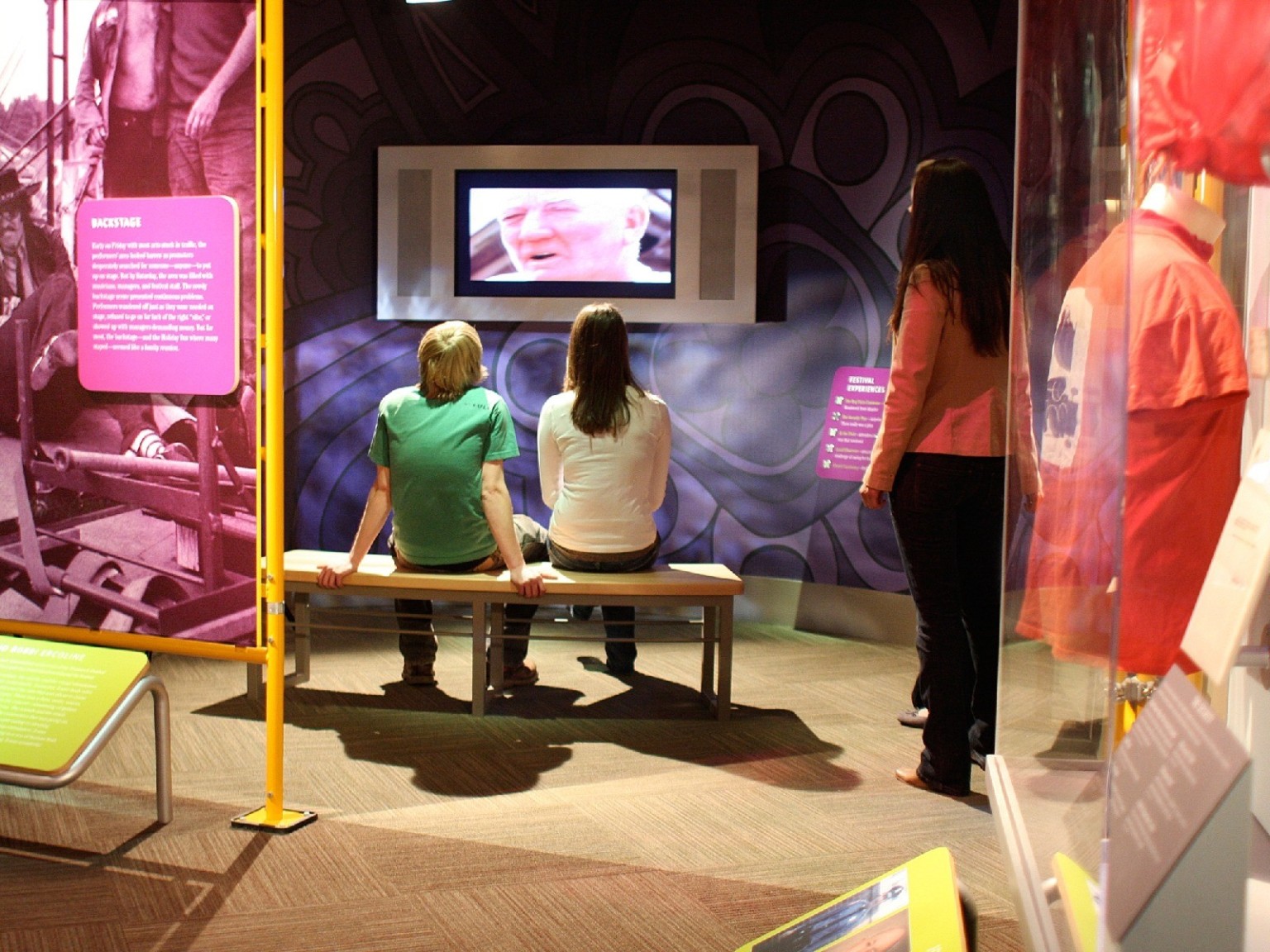 Two people sit in a side room of the museum watching a video on a screen built into a wall with abstract design