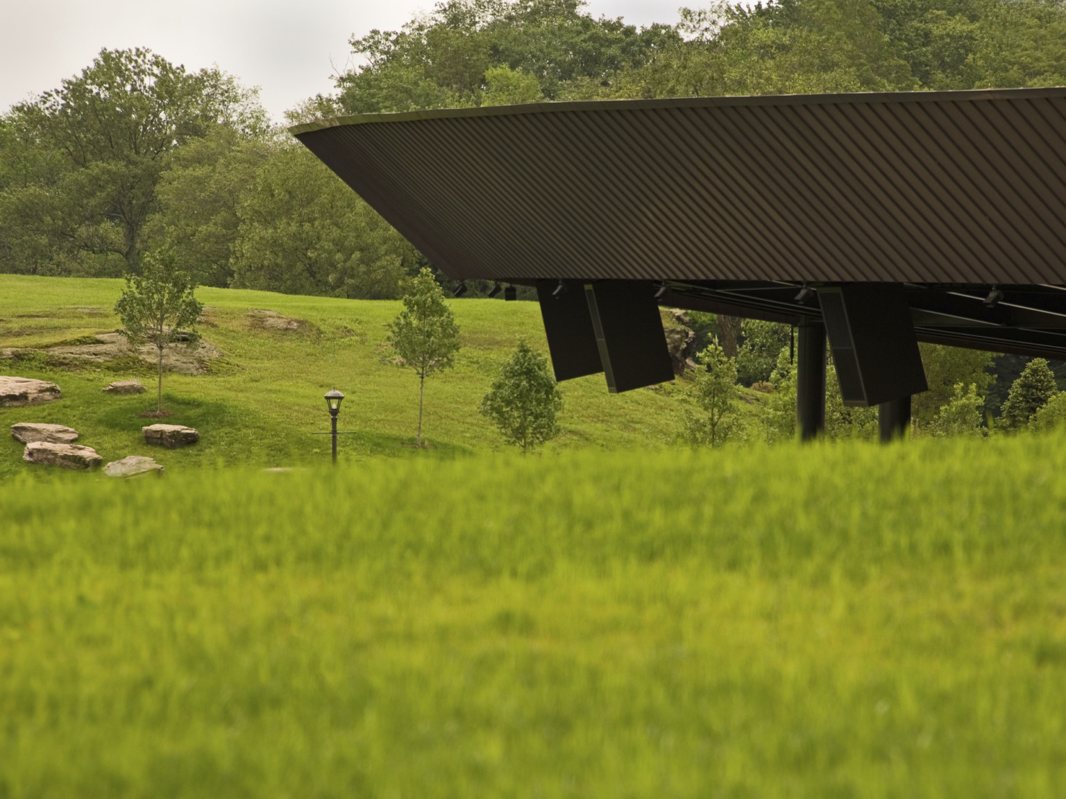 Fascia of amphitheater is visible above green grassy hill; beyond the building is another hill cover with trees; Bethel Woods Center for the Arts
