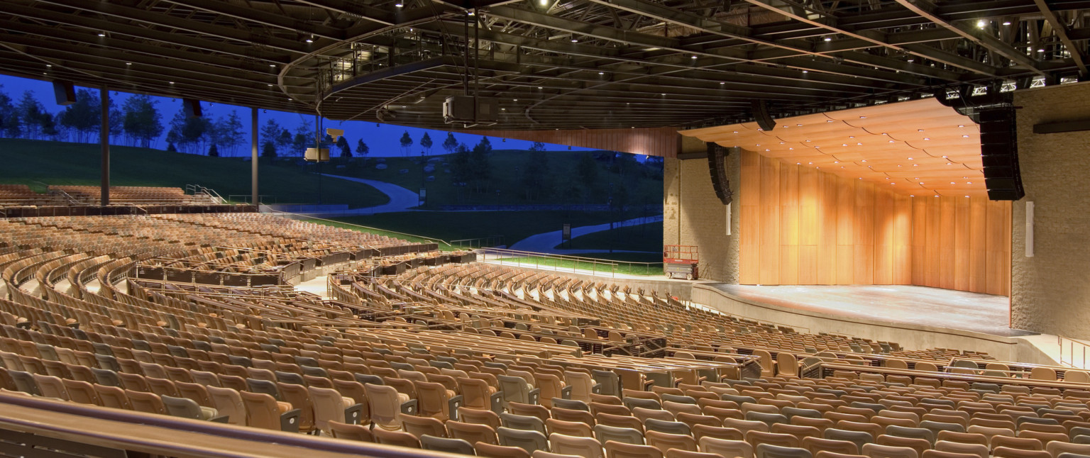 Light brown seats under black amphitheater roof facing wood paneled stage with rounded panel ceiling and recessed lights