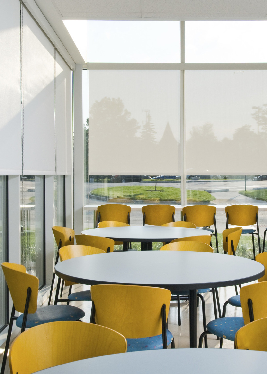 Round tables with multicolored chairs in room with floor to ceiling windows, translucent white roller shades partially drawn