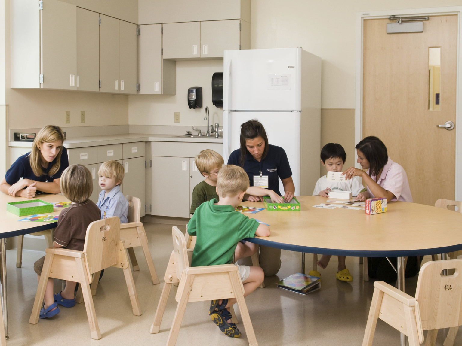 Wood children's chairs and round tables in cream colored room with refrigerator and sink in counter with cupboards