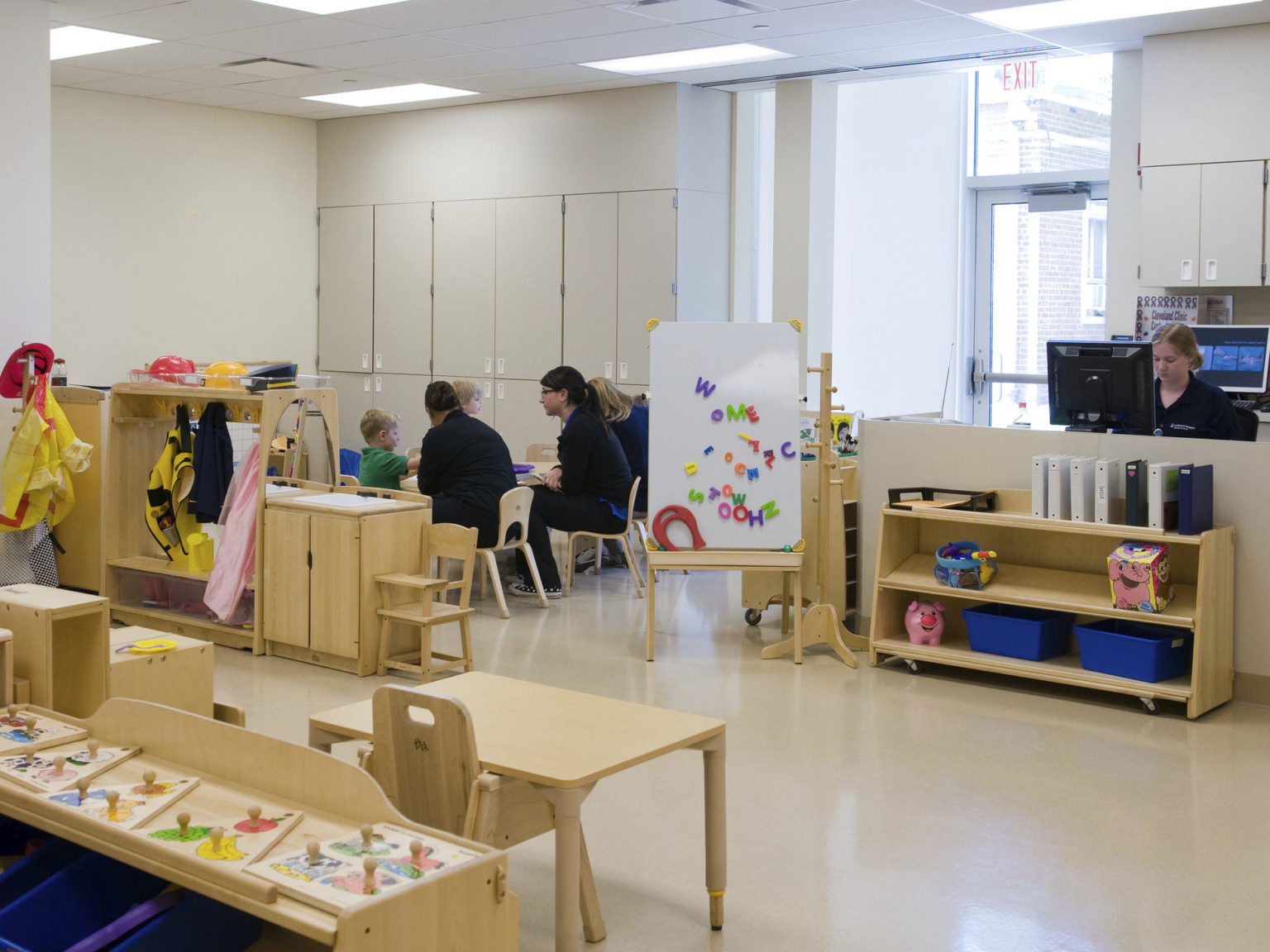 Playroom with wooden tables and shelves. Cream cupboards along the back left wall separated from glass door by white wall