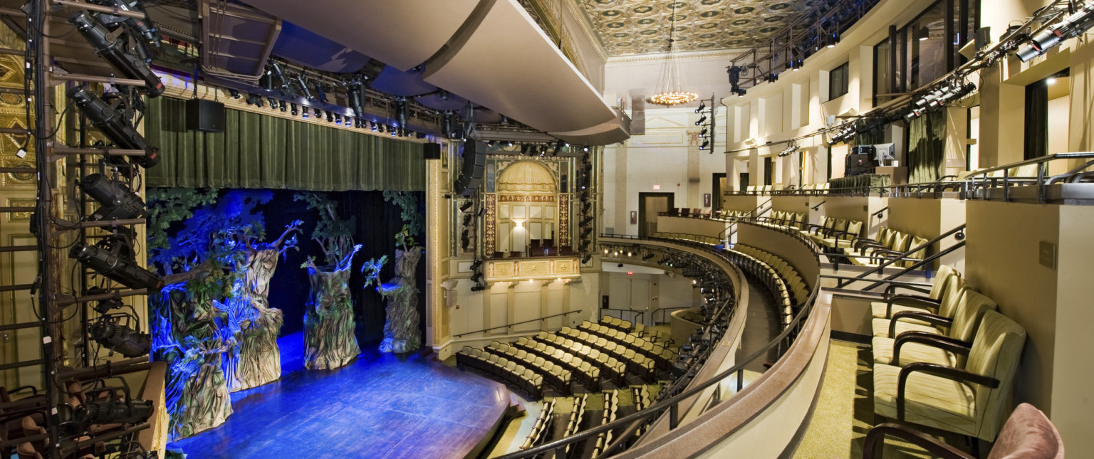Multiple balconies in a theater with yellow seats and dark details, box seat center with arch overhang and gold details