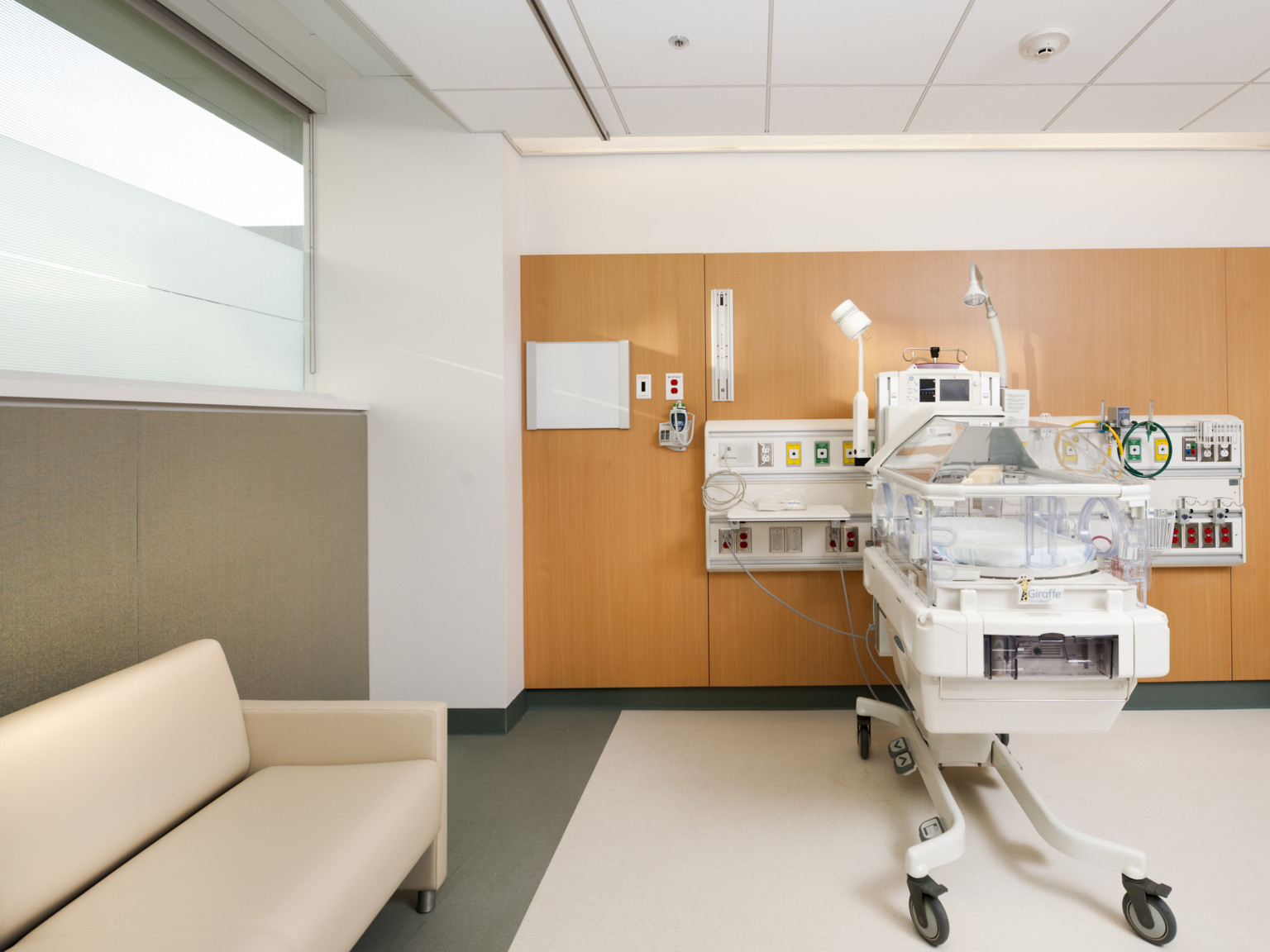 Incubator for babies sits to the right with wood back wall. Cream colored couch sits near by to the left against beige wall