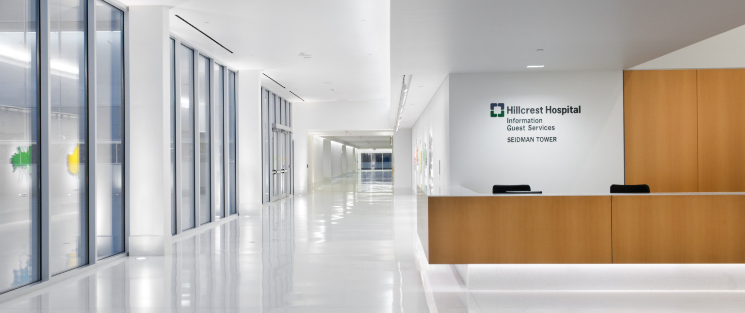 White hallway with wood accent reception desk, wall above reads Hillcrest Hospital Information Guest Services Seidman Tower