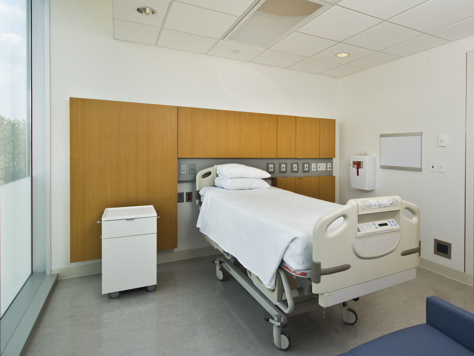Hospital bed in the center of a white room with wood accent behind. There is grey tile flooring, and floor to ceiling window