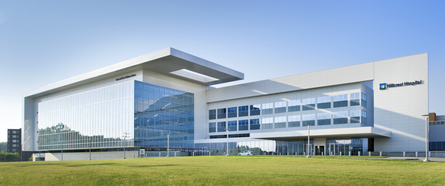 Front of hospital viewed from across the grass. Multistory windowed outcrop wraps around left section with overhang above