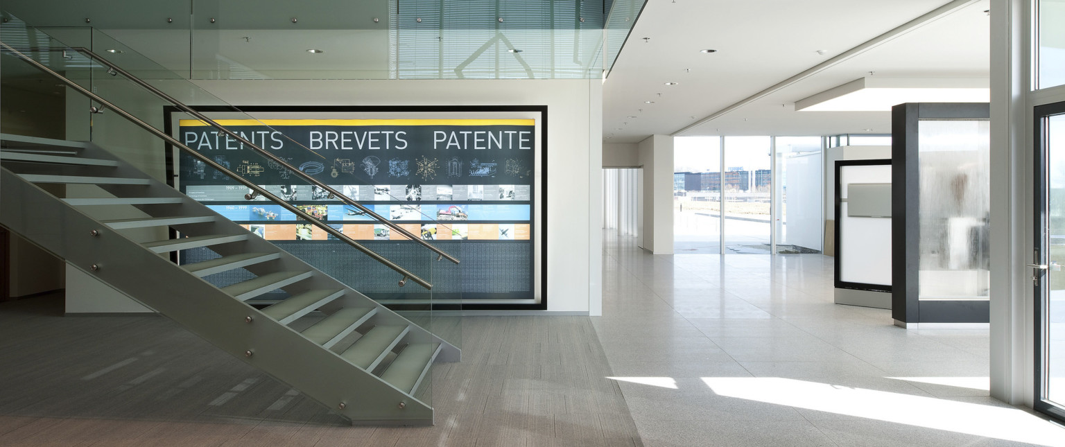 White hallway with floor to ceiling windows, right. Floating stairs with glass bannister, left. Back, infographic of patents