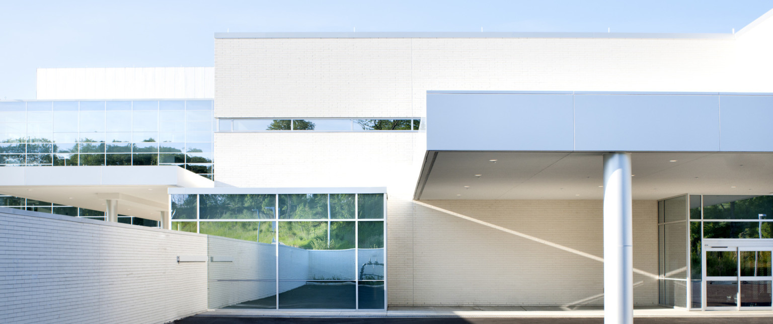 White stone building. Outcrop with floor to ceiling windows left with stone wall. Grey metal pillar with overhang right