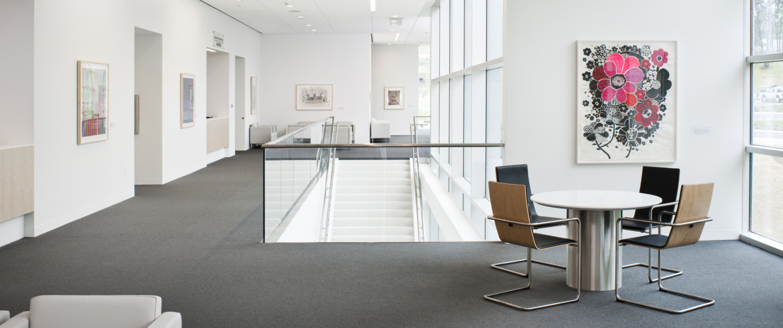 White room with multiple seating areas and stair case center coming from below next to window wall. Art is framed on wall