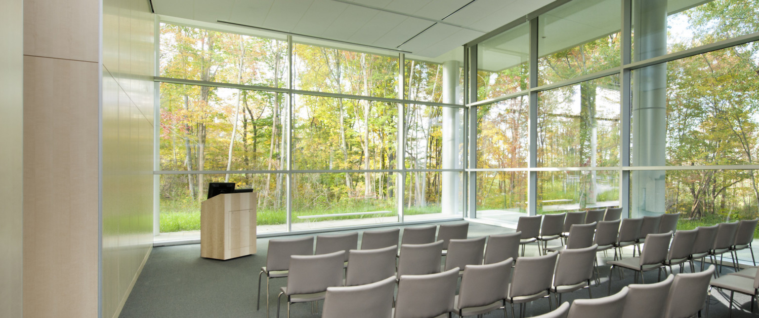 Auditorium with grey floor, wood left wall, and floor to ceiling windows on two walls with ground level view of trees.