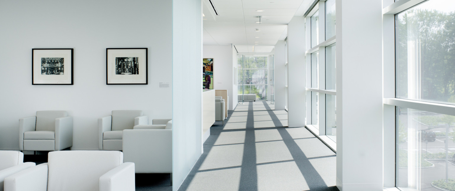 Hallway with floor to ceiling windows right and at end. Left divided into sections by white wall, each has seating areas