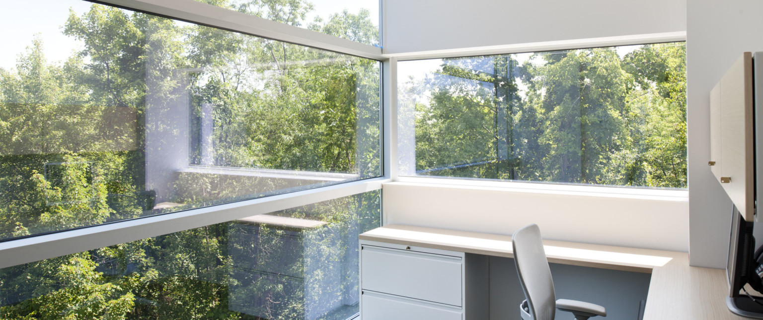 Corner view of office with L shaped desk and grey chair. Left is floor to ceiling window, back wall is white with wide window