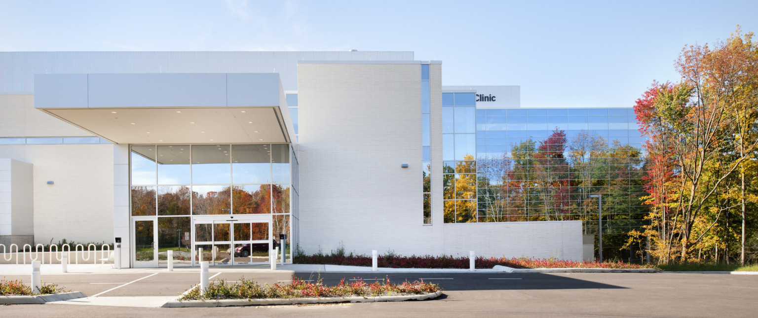 Entry with double height windows with white overhang extending over driveway. White wall right with window wall beyond