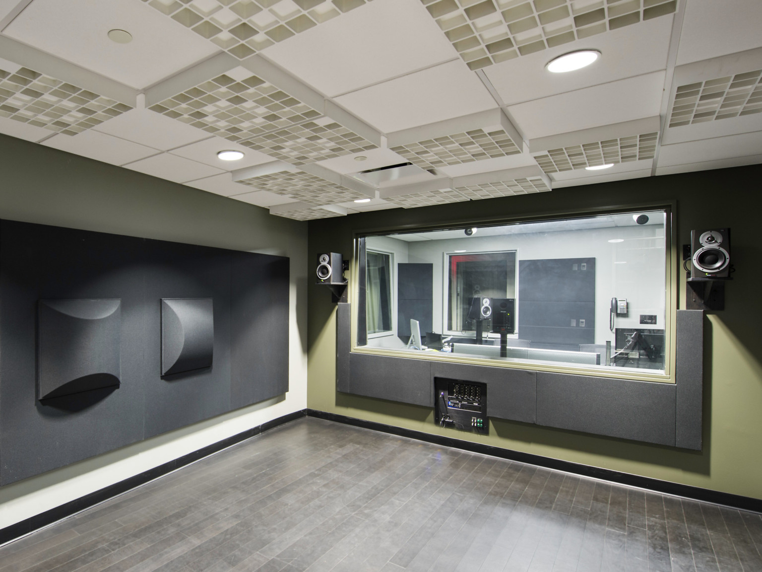 Green room with black acoustic wall panels and cream colored panels on the ceiling, and a window looks into control room