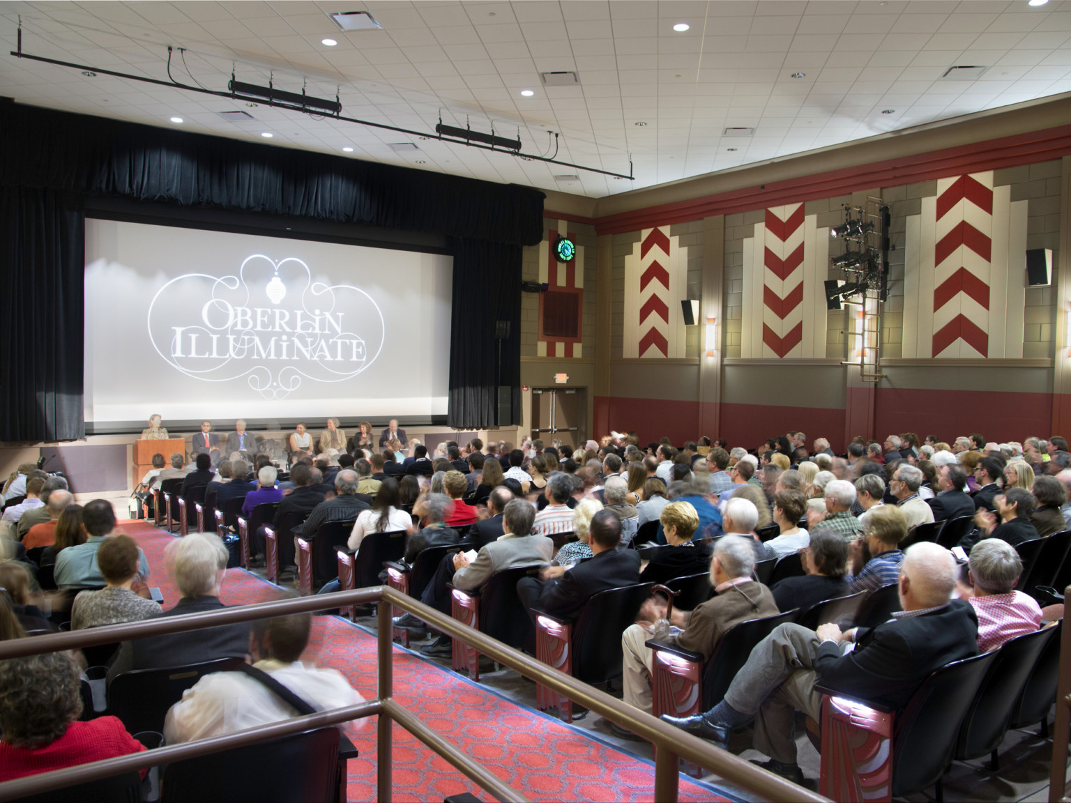 Audience sits in movie theater with geometric red carpeting and geometric tiled wall pattern