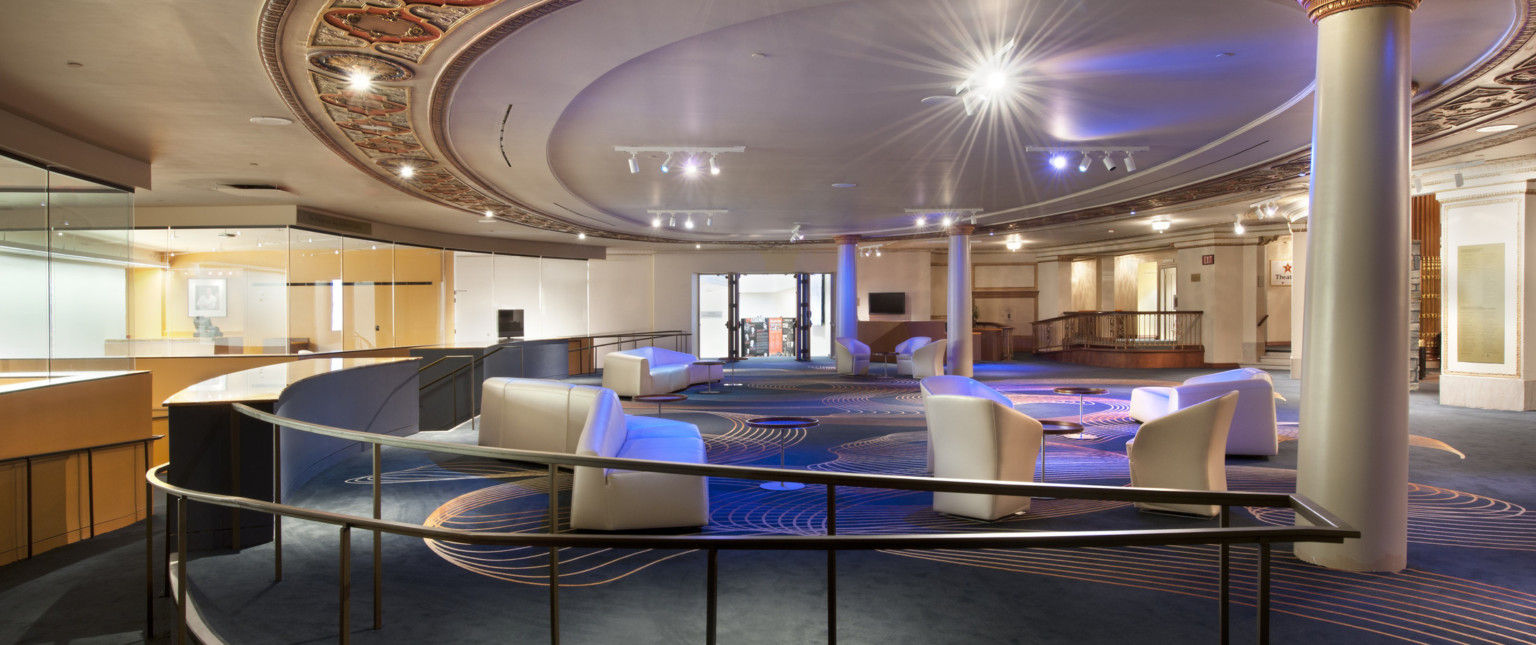 Lobby with dark colored carpet with gold organic design. White angled seats through out the room below a round ceiling detail