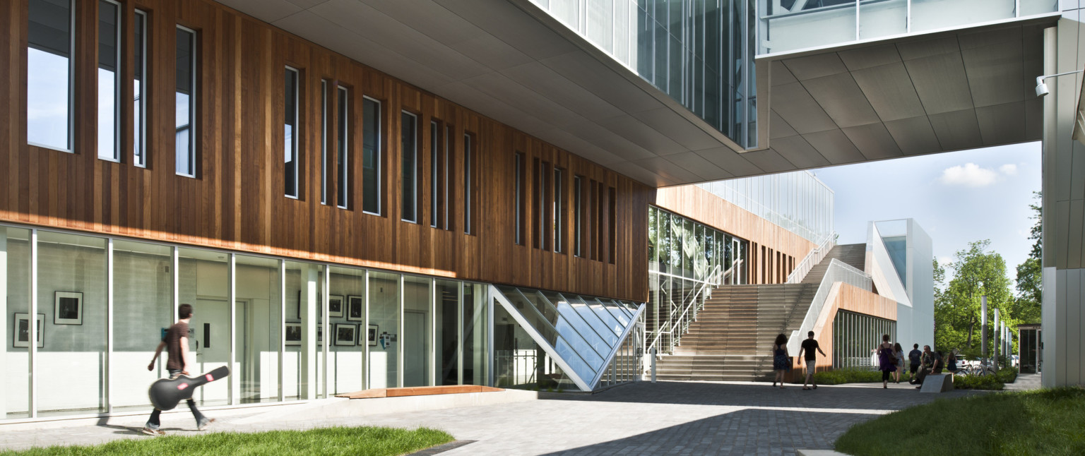 Partially covered walkway between buildings, building at left has first floor window walls below wood panels