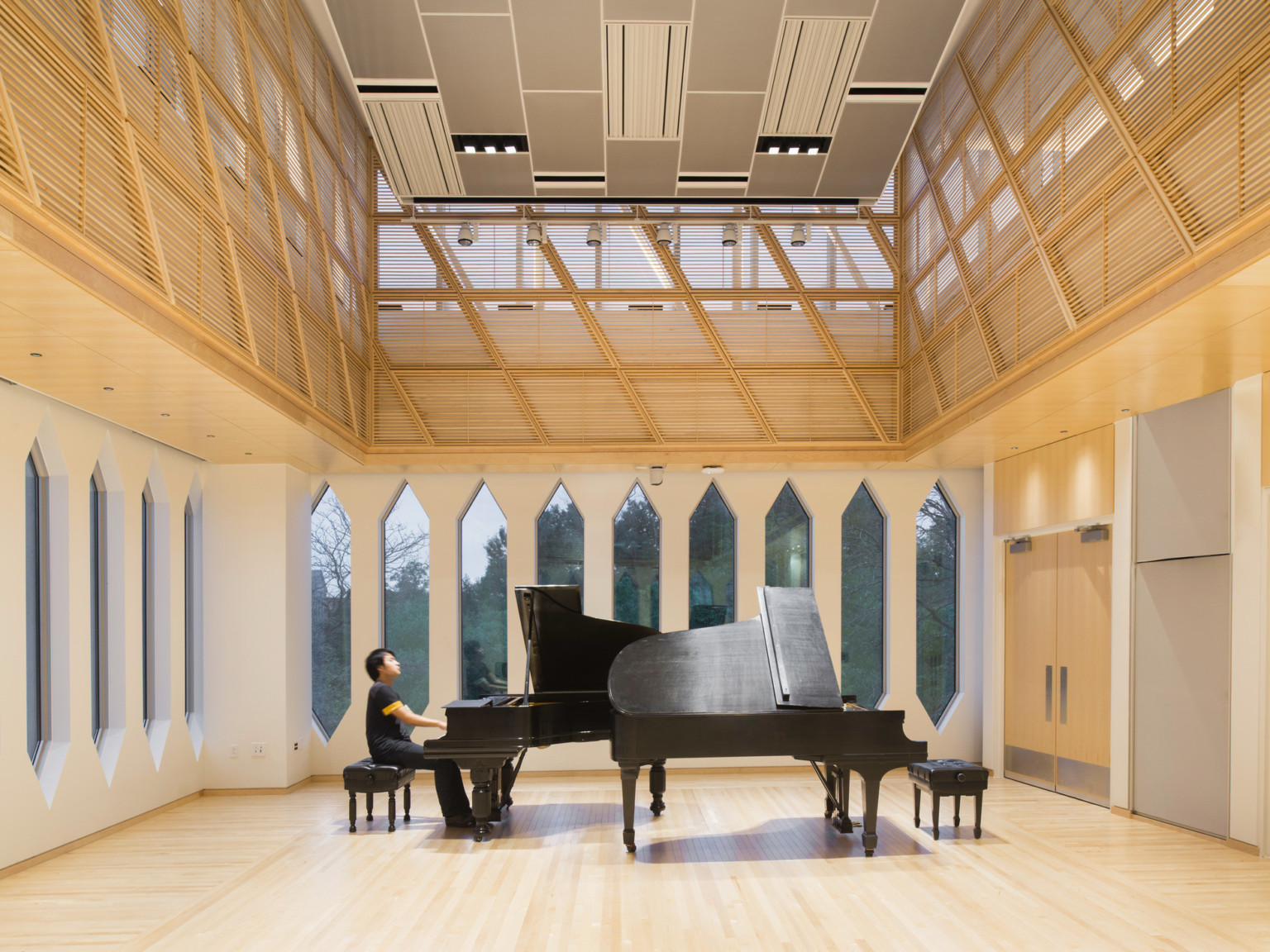 Corner room with white walls and thin rectangular windows with pointed tops and bottoms. A piano, center, on light wood floor