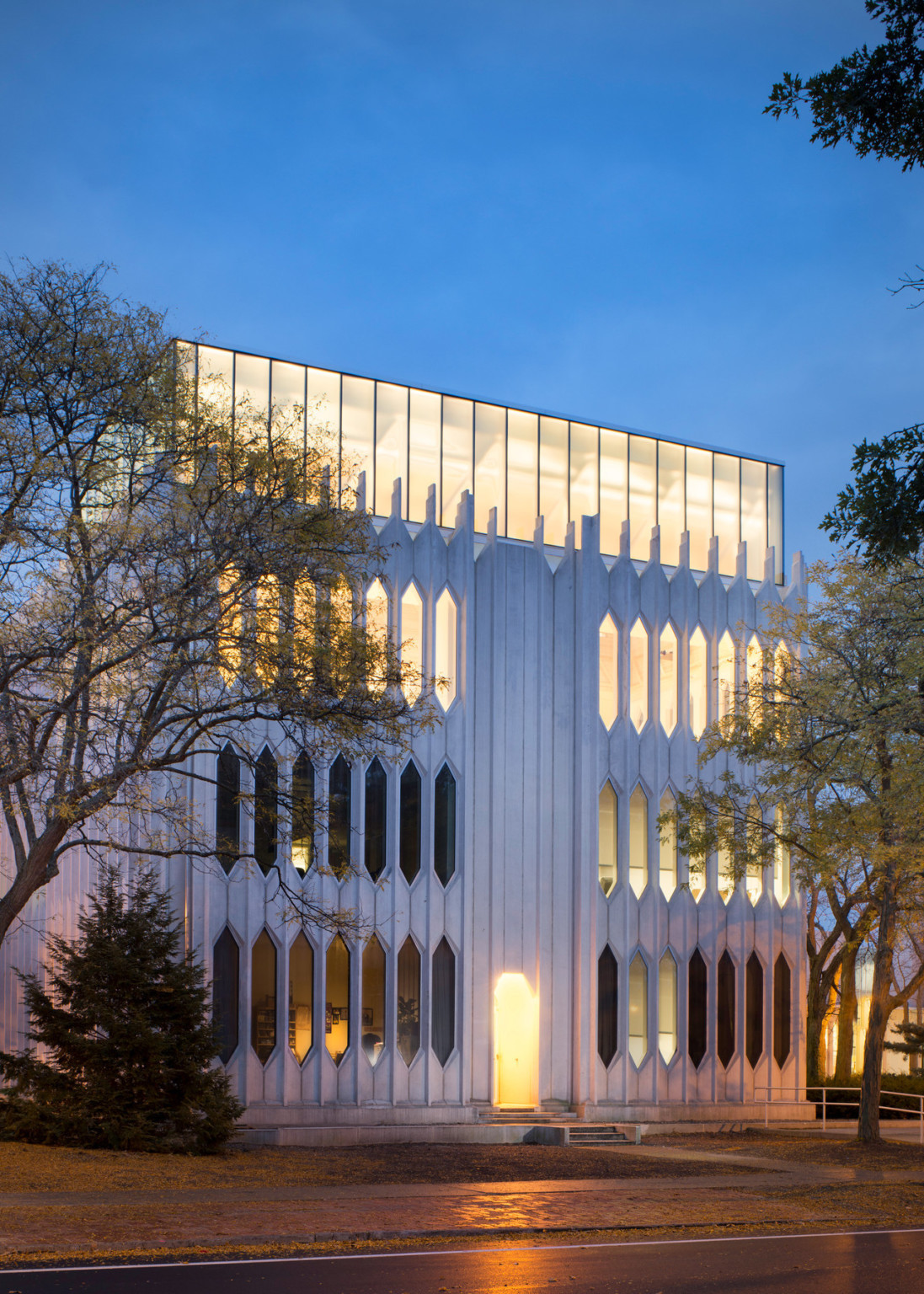 Building illuminated from within, glass top floor extends above white exterior on lower levels with thin pointed windows