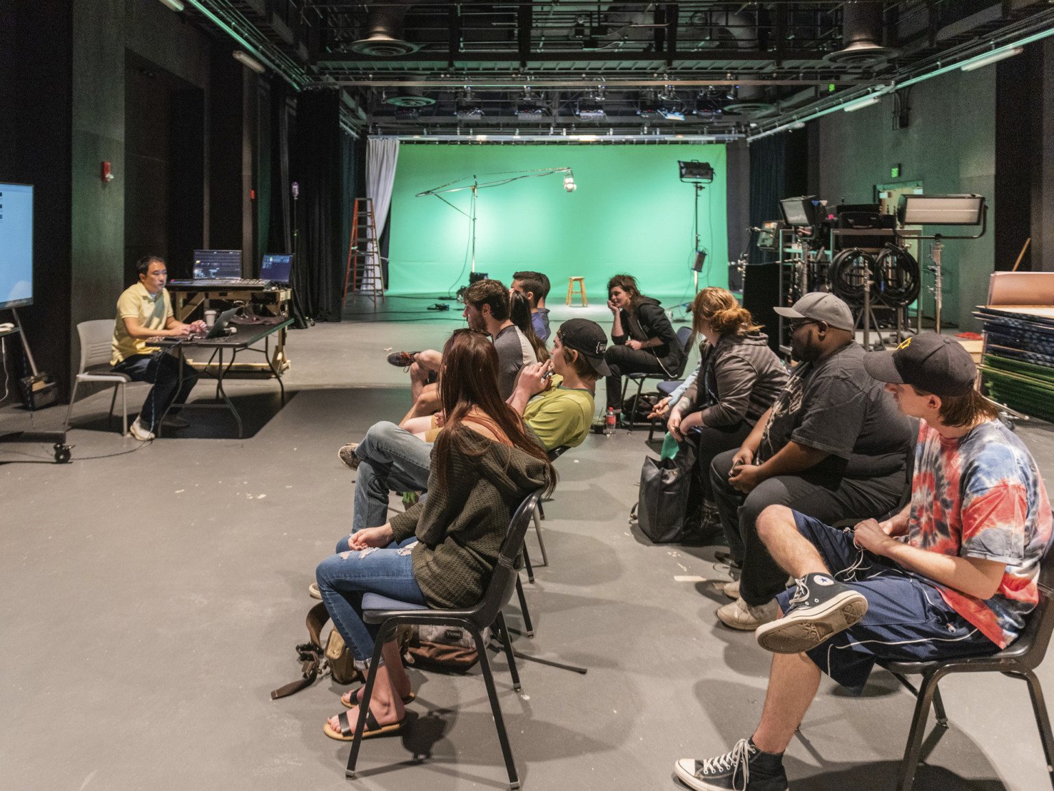 Students sit in chairs in a long dark room, at the end of which is a green space that can be separated by a curtain