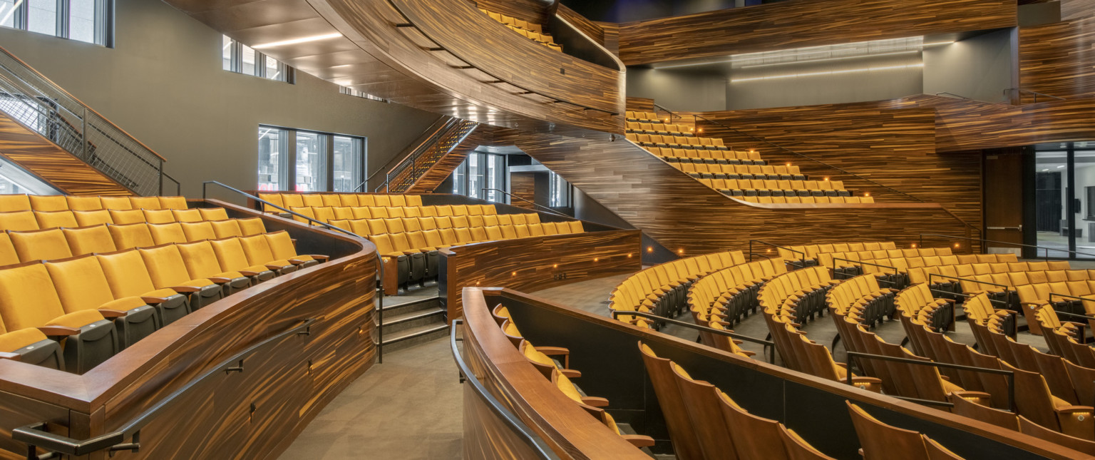 Stadium style yellow seating and abstract stripe stained wood details, balcony, and bannisters with recessed aisle lighting