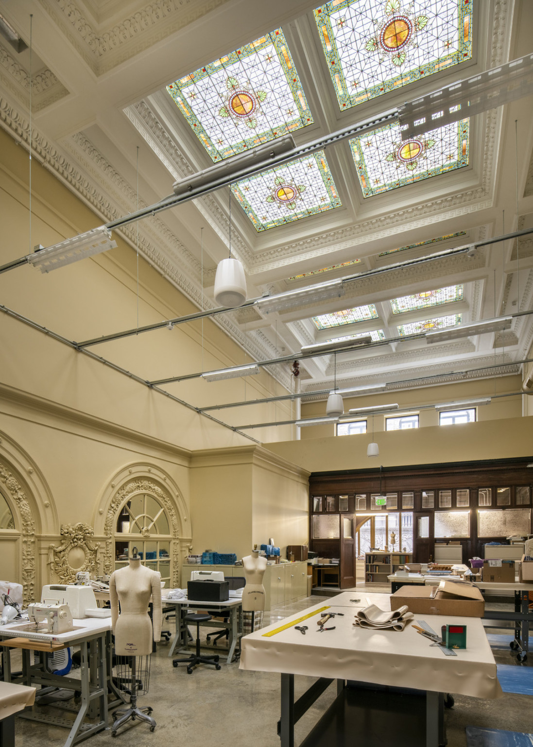 Green and yellow skylights above lighting system on rafters shed light on long room with sewing machines and arched mirrors